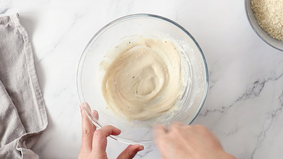 Oat milk, flour, and seasonings being mixed together in a glass bowl to make a thin batter.