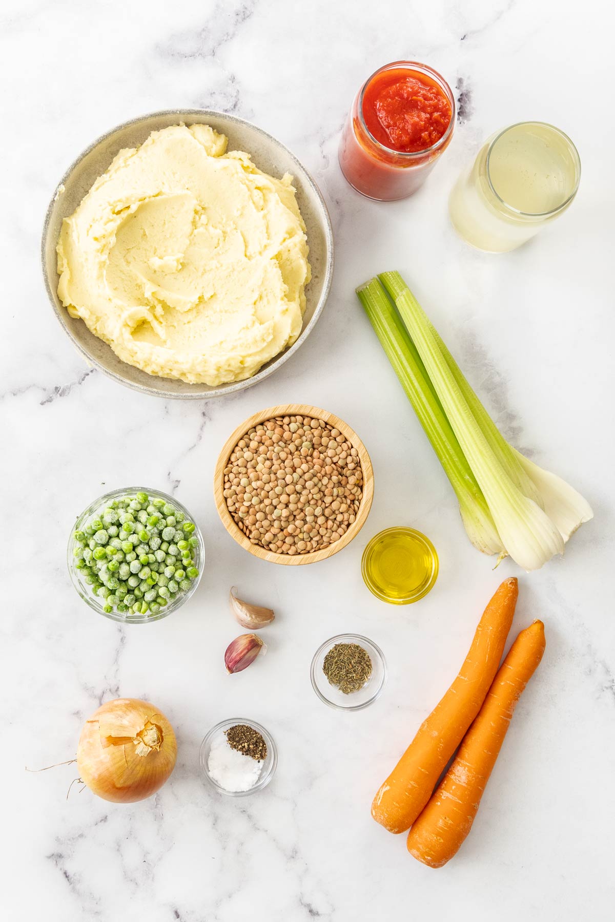 Overhead photo with mashed potatoes, lentils, frozen peas, onion, carrots, celery, canned tomatoes, vegetable broth, olive oil, and spices on a marble background.