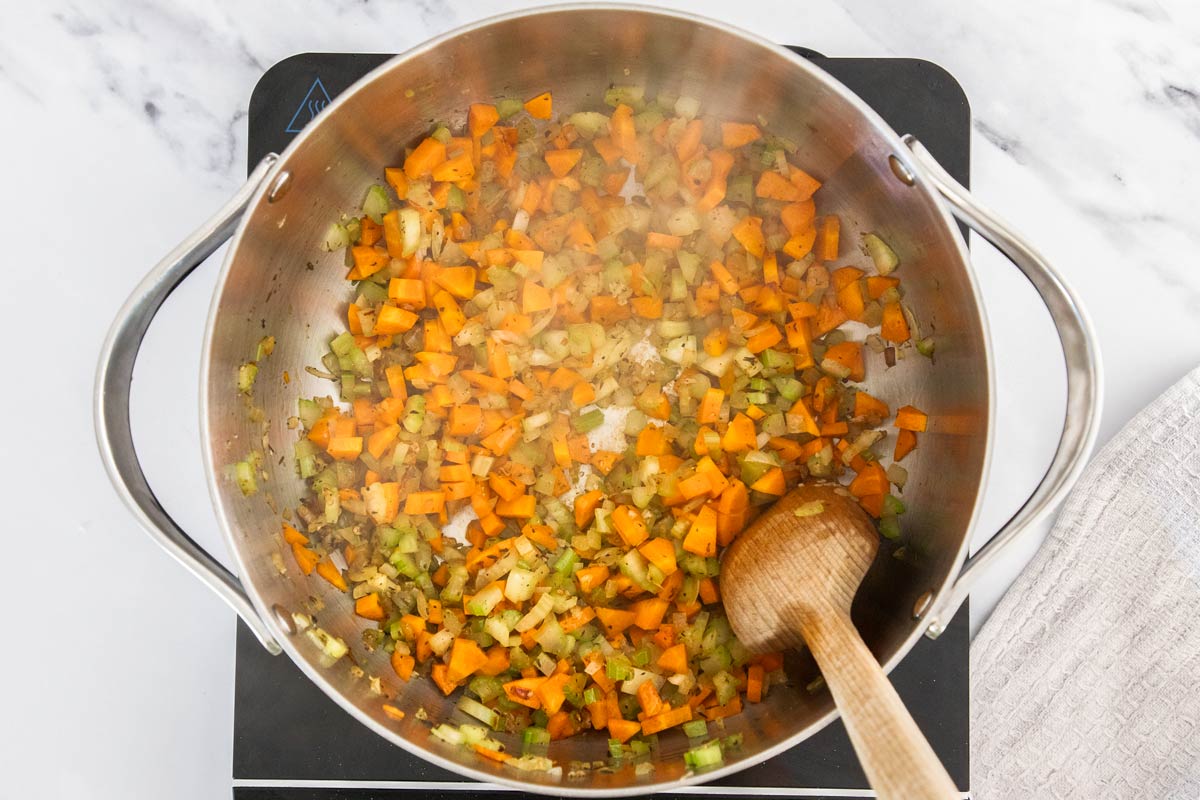 Overhead photo of carrots and celery being sautéd with onions in a stainless steel saucepan.