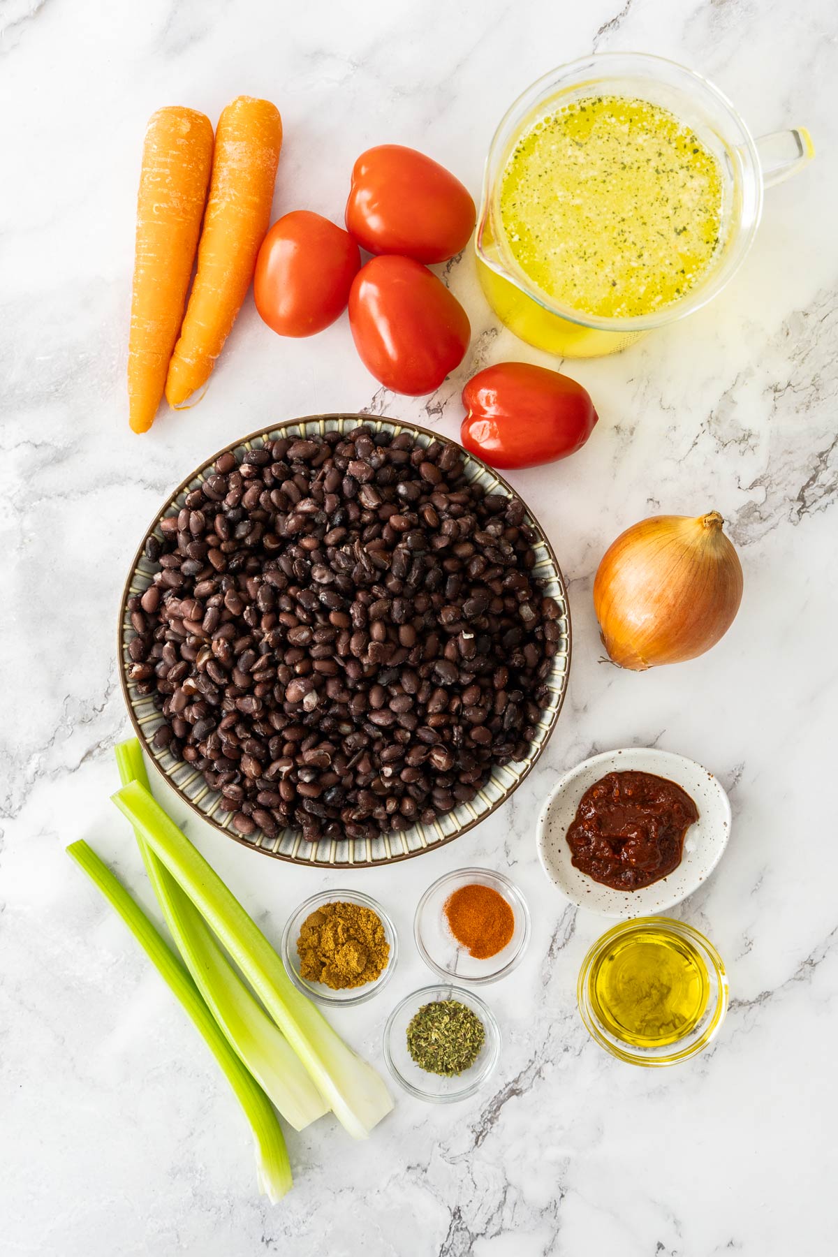 Black beans, tomatoes, carrots, celery, onion, garlic, chipotle, pepper, vegetable broth, spices, and olive oil on a marble background.