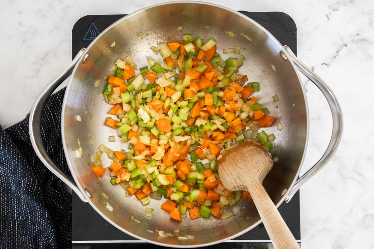 Overhead photo of onions, celery, carrots, and garlic being sautéed in a stainless steel soup pot.