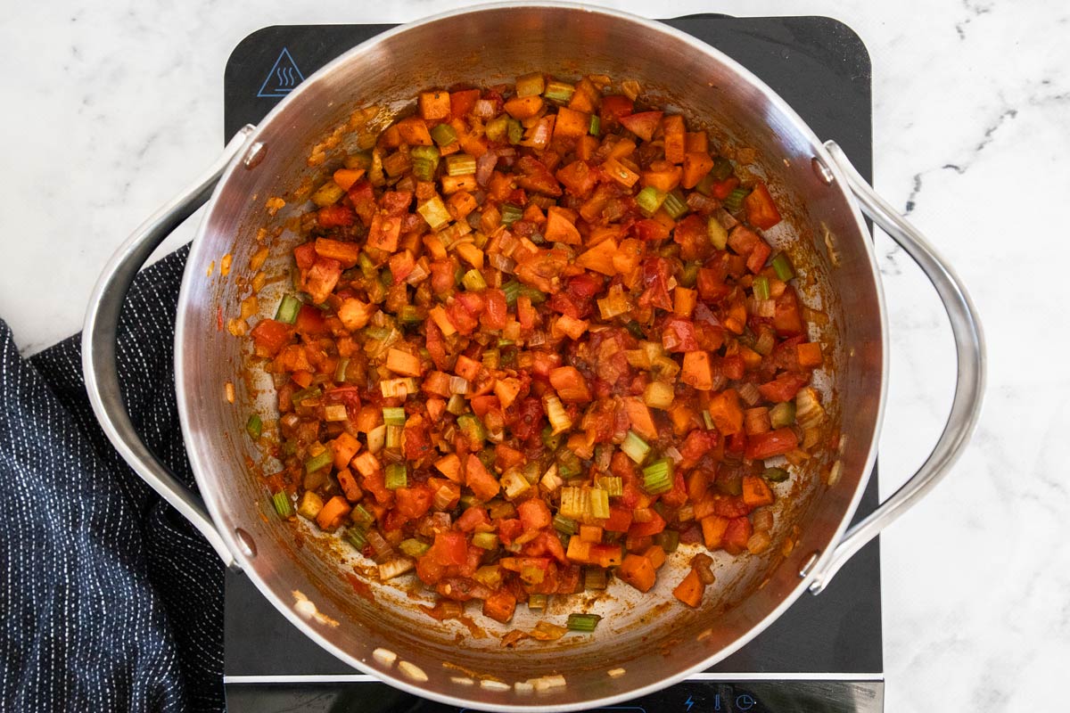 Overhead photo of vegetables cooking in a stainless steel soup pot, with tomatoes and spices added in.