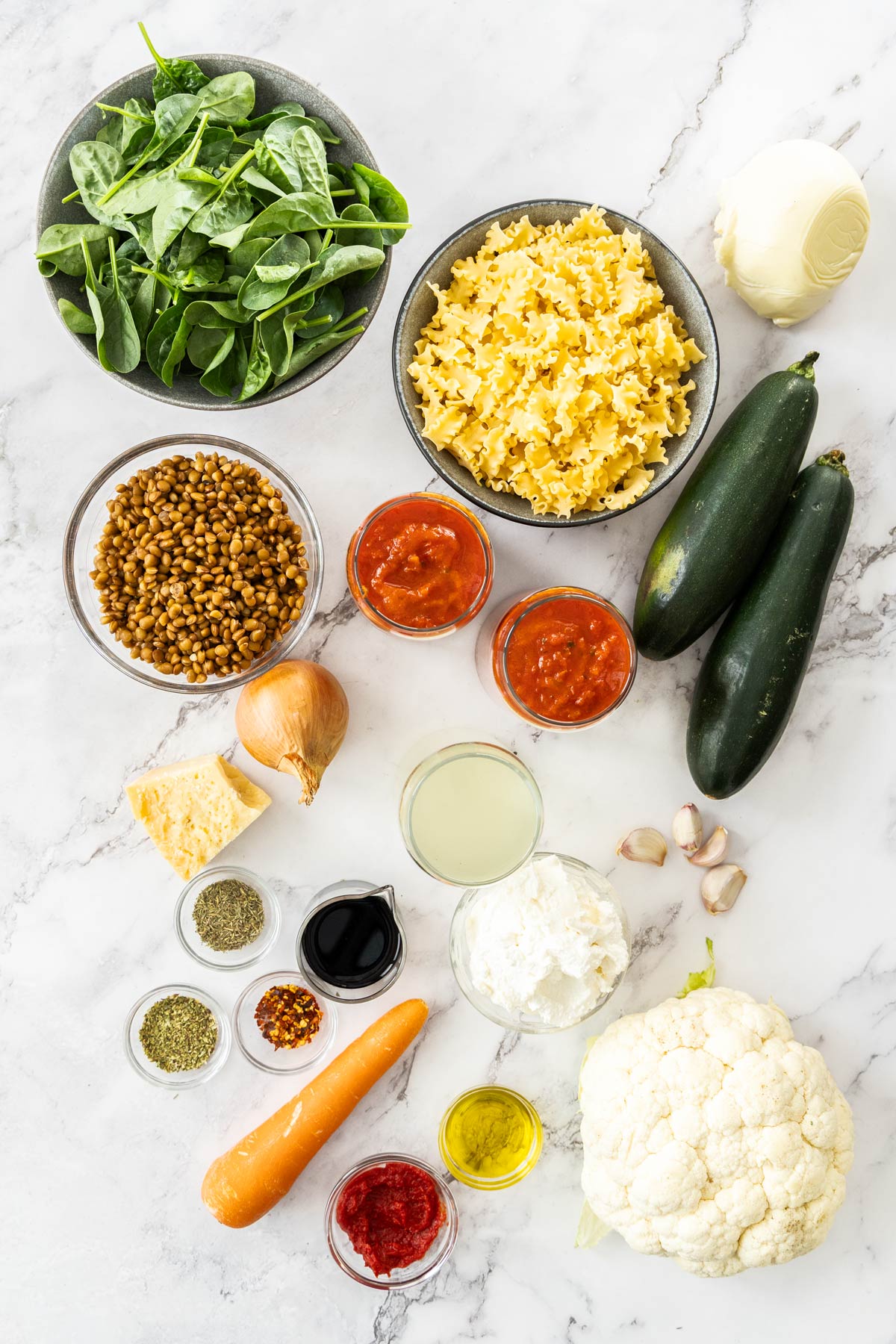 Spinach, pasta, lentils, mozzarella, ricotta, onion, tomato sauce, zucchini, cauliflower, carrot, vegetable broth, tomato paste, and spices on a marble background.