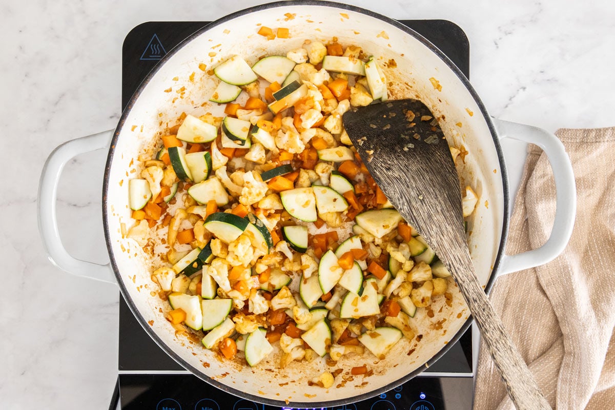 Overhead photo of onions, zucchini, carrots, and cauliflower florets being sautéd in a white ceramic skillet.