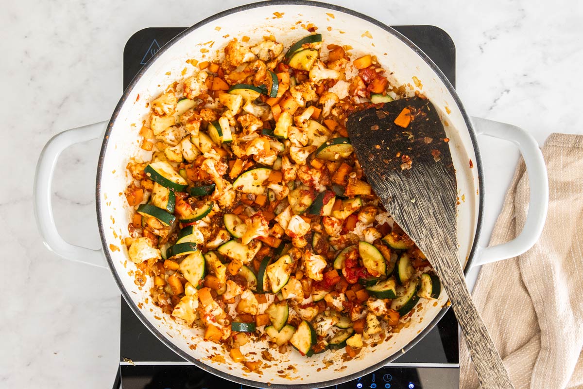 Overhead photo of vegetables being stirred in a white ceramic skillet with tomato paste and spices added in.