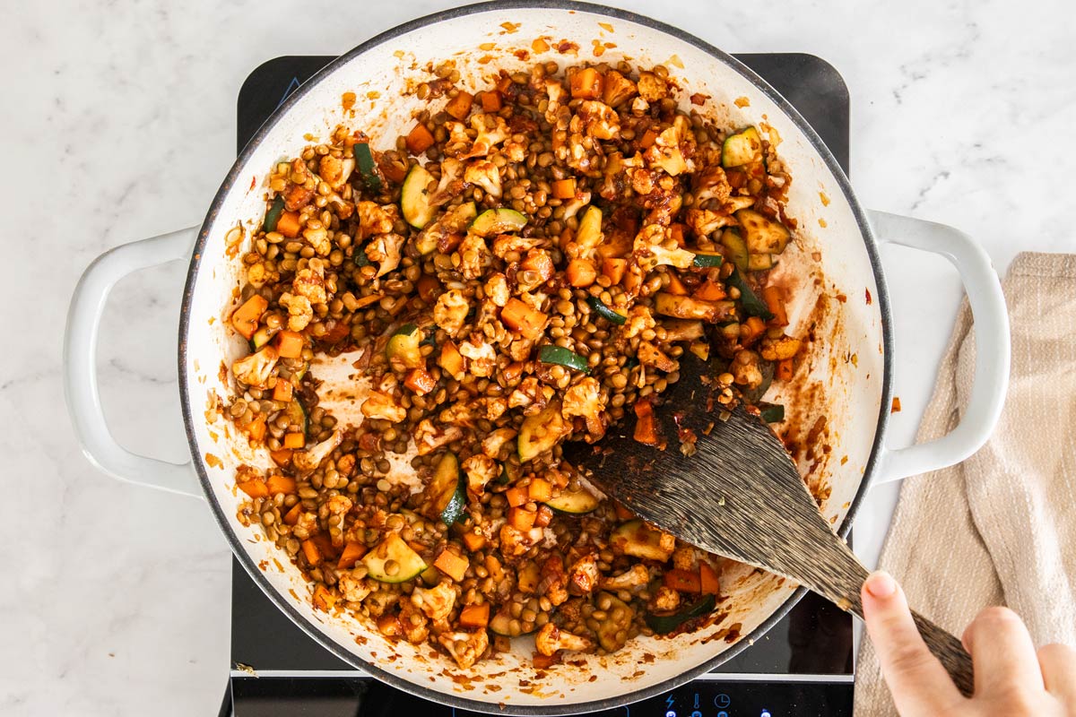 Overhead photo of vegetables being cooked in a white heavy-bottomed skillet with lentils being stirred in.