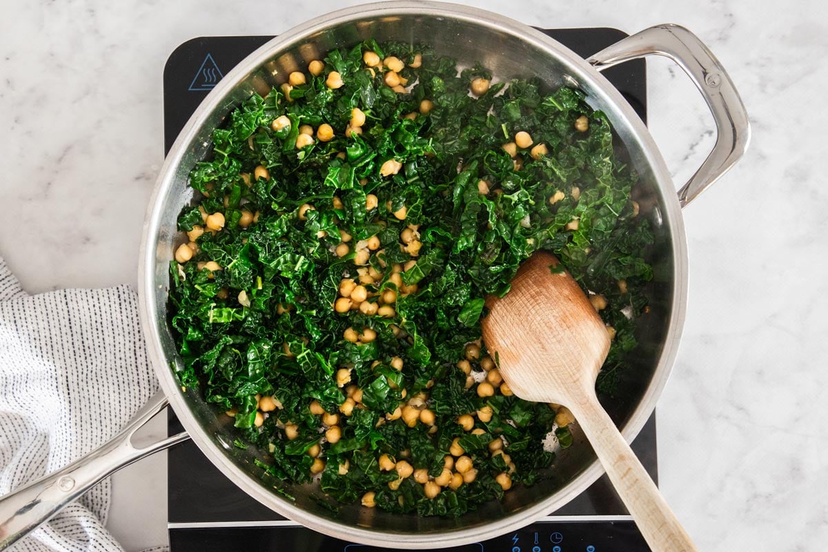 Overhead photo of chickpeas and kale being sautéd in a stainless steel skillet.