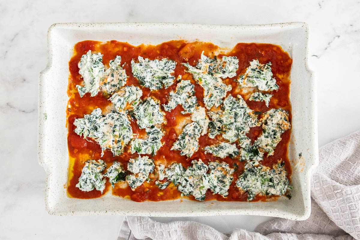 A white lasagna pan with marinara sauce on the bottom and dollops of a spinach and ricotta mixture over it.