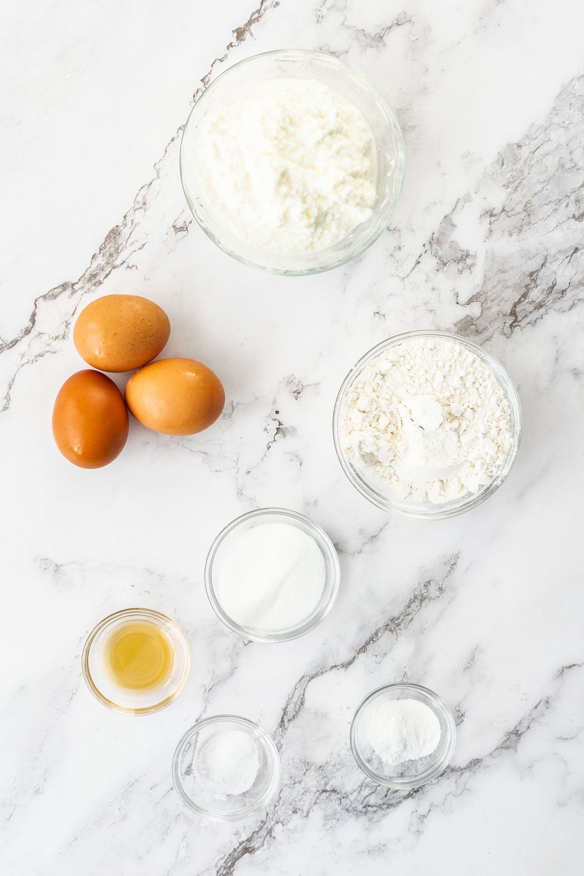 Eggs, cottage cheese, flour, melted butter, sugar, and baking powder on a marble background