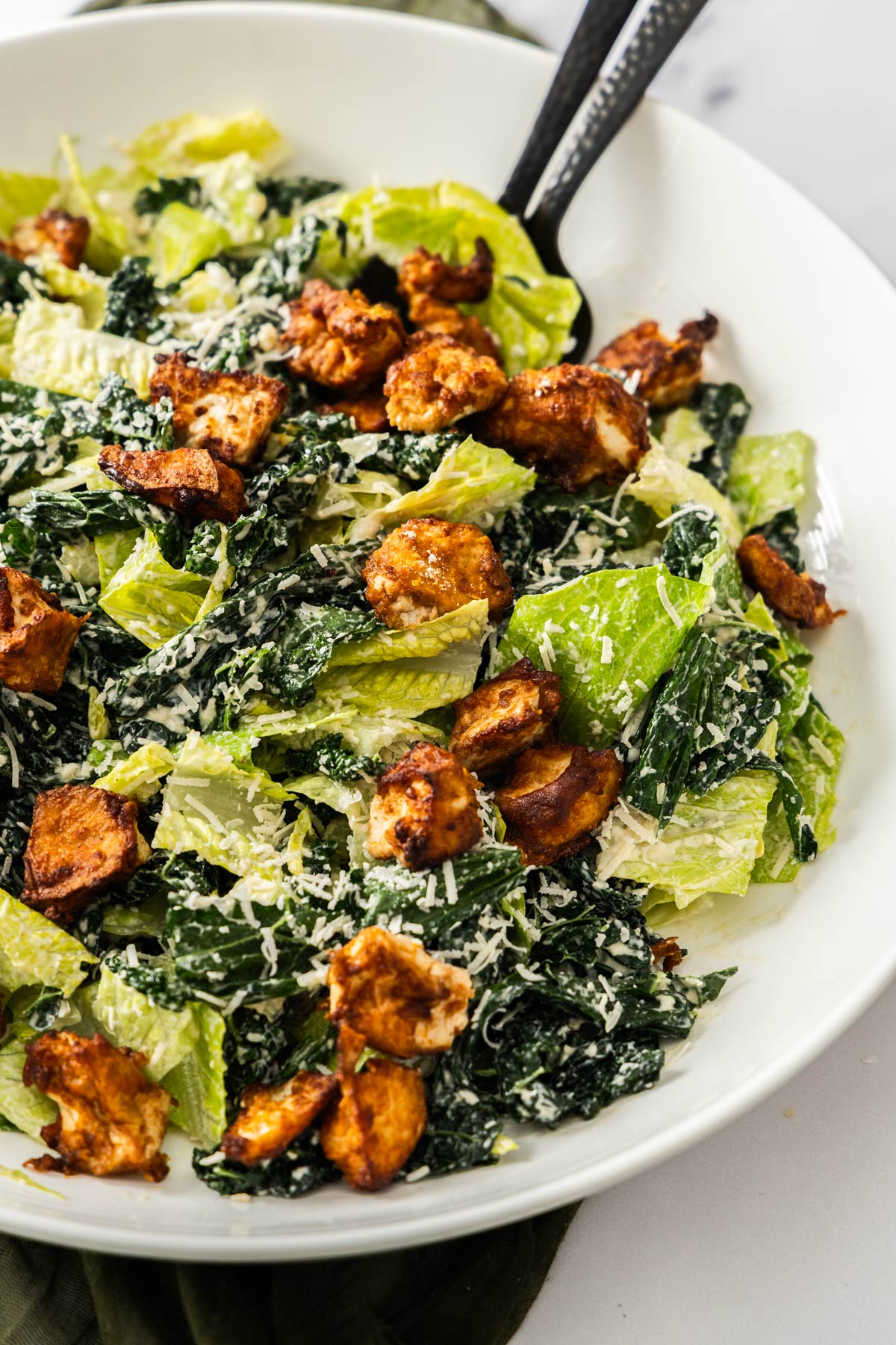 Close up shot of a half kale half romaine caesar salad topped with crispy tofu in a white salad bowl