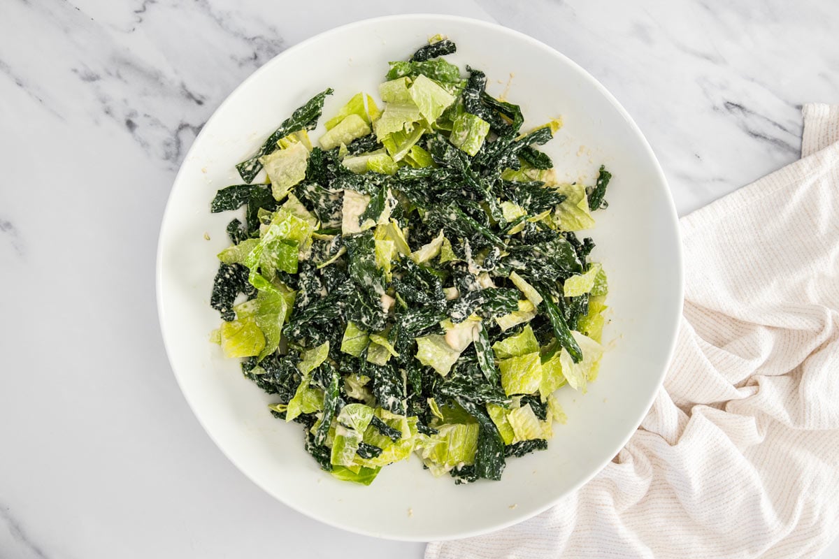 Kale and romaine caesar salad being mixed in a white salad bowl