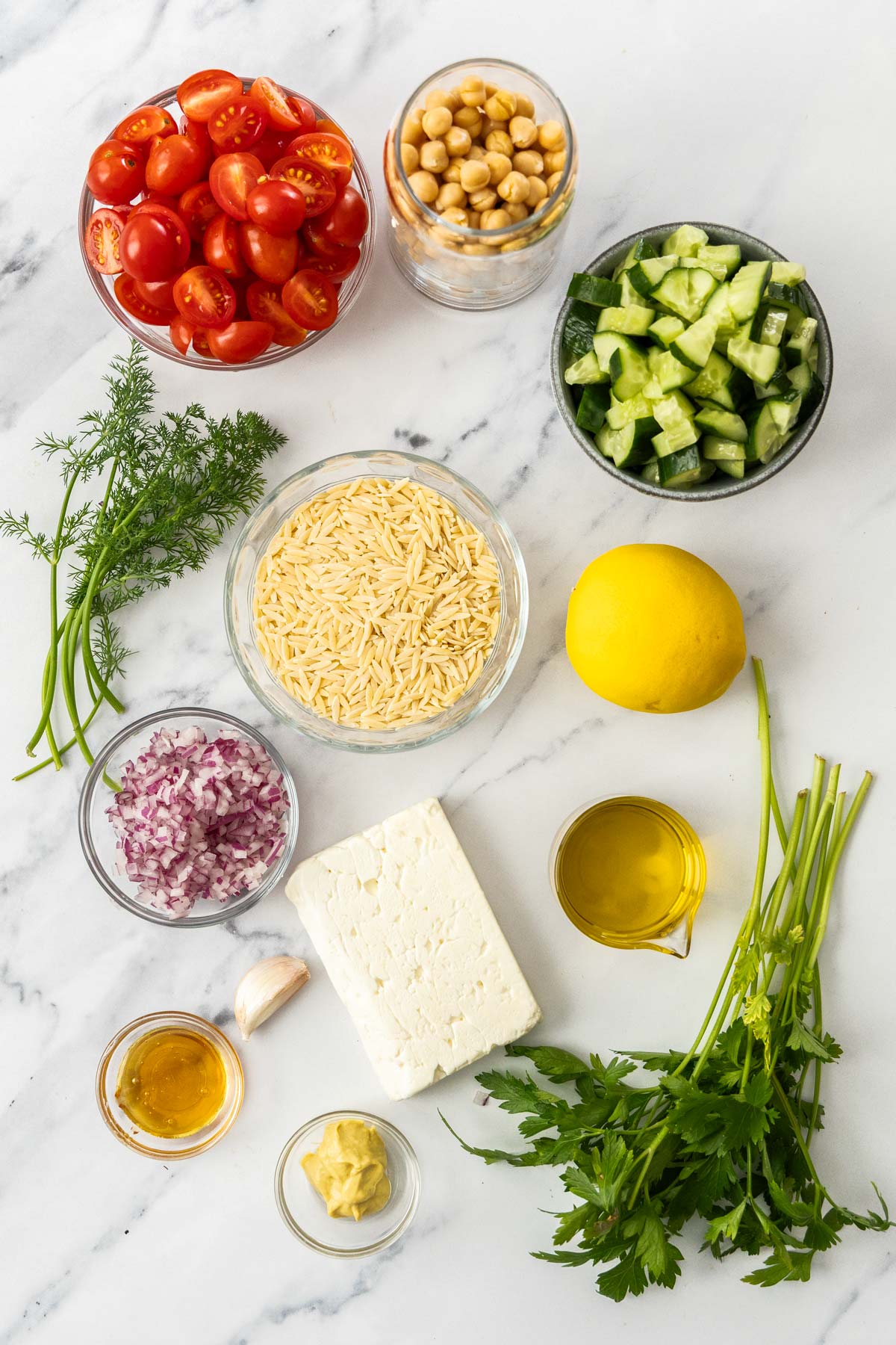 Overhead photo of dried orzo, diced cucumber, sliced cherry tomatoes, chickpeas, diced red onion, a block of feta, fresh herbs, dijon mustard, honey, garlic, and olive oil on a marble background.