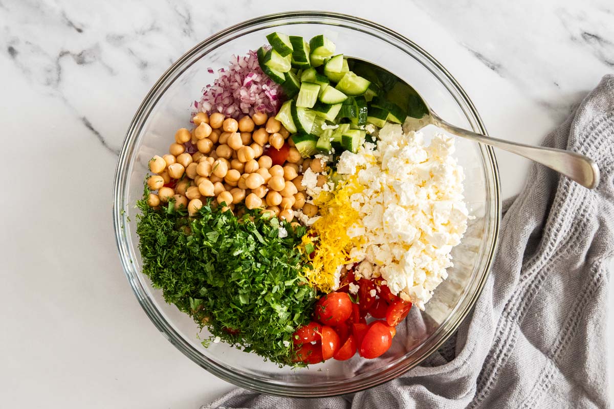 Overhead photo with chopped cucumber, tomatoes, herbs, chickpeas, and crumbled feta in a glass bowl